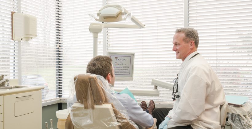 In a brightly lit dental office, a male dentist in a white lab coat discusses treatment with a male patient seated in a dental chair, under a backdrop featuring a computer monitor displaying information about Botox and dermal fillers.