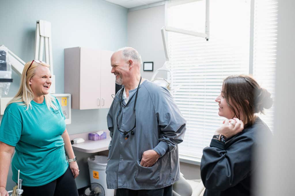 A group of dental professionals are having a cheerful conversation in a dental office. The team includes a dentist and two dental assistants, all smiling and engaged. The office setting is equipped with standard dental equipment, including a dental chair, cabinets, and a light source near the window. The atmosphere appears friendly and professional.