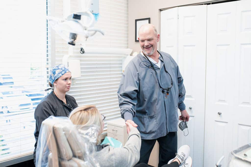 A dental professional wearing a gray jacket shakes hands with a patient seated in a dental chair, while another dental professional in black attire and a blue patterned surgical cap sits nearby.