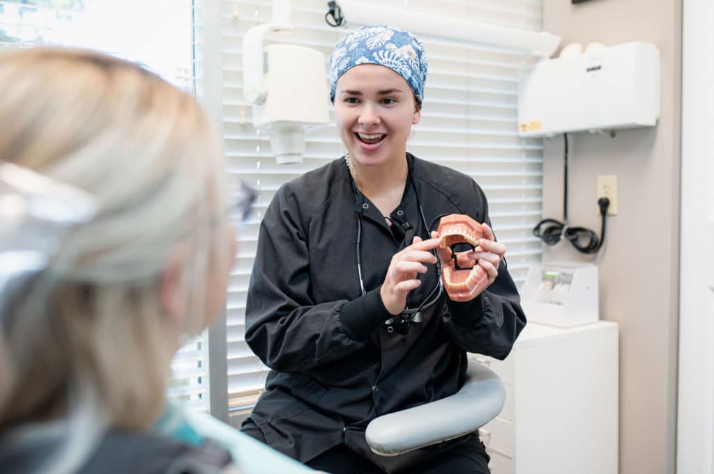 A dental professional wearing a blue patterned surgical cap and black attire explains dental health to a patient by using a model of a mouth.