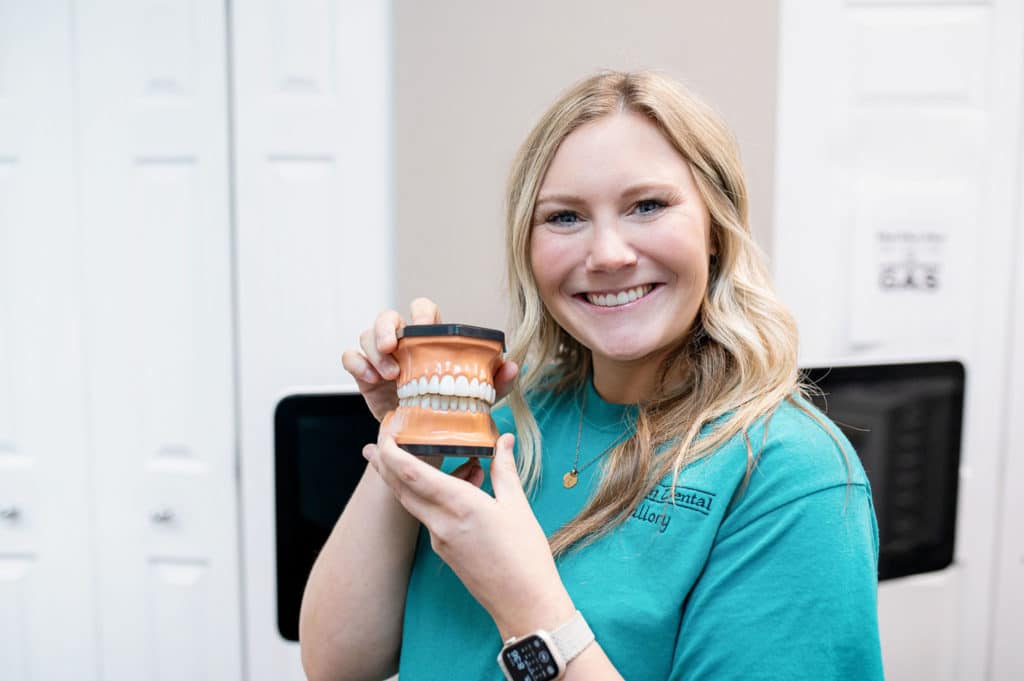 A smiling dental professional wearing a turquoise shirt holds up a dental model, displaying a set of teeth. She stands in a dental office, showing the model to the camera.