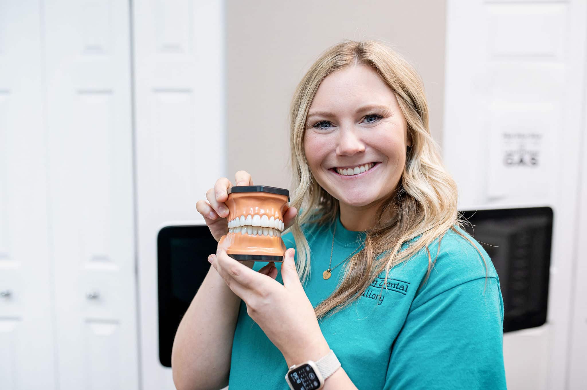 A smiling dental professional wearing a turquoise shirt holds up a dental model, displaying a set of teeth. She stands in a dental office, showing the model to the camera.