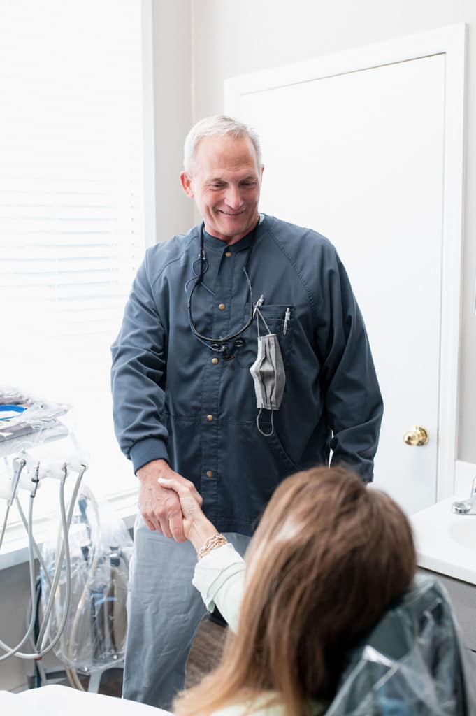 A dentist warmly shaking hands with a patient seated in a dental chair, both smiling, in a bright dental clinic room.