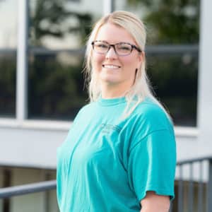 A woman with long blonde hair and glasses smiles while posing outdoors. She is wearing a teal shirt with text on the chest and standing in front of a building with large windows.