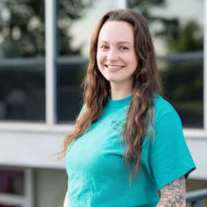 A woman with long, wavy brown hair smiles while posing outdoors. She is wearing a teal T-shirt and standing in front of a building with large windows. She has a tattoo visible on her left forearm.