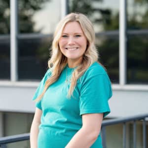 A woman with long blonde hair smiles while posing outdoors. She is wearing a teal shirt and standing in front of a building with large windows.