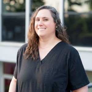 A woman with curly brown hair smiles while posing outdoors. She is wearing a black scrub top and standing in front of a building with large windows.