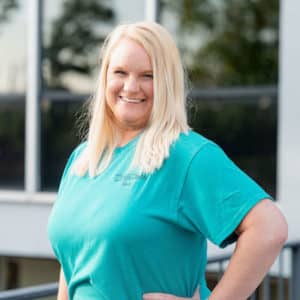 A woman with long blonde hair smiles while posing outdoors. She is wearing a teal shirt and standing in front of a building with large windows.