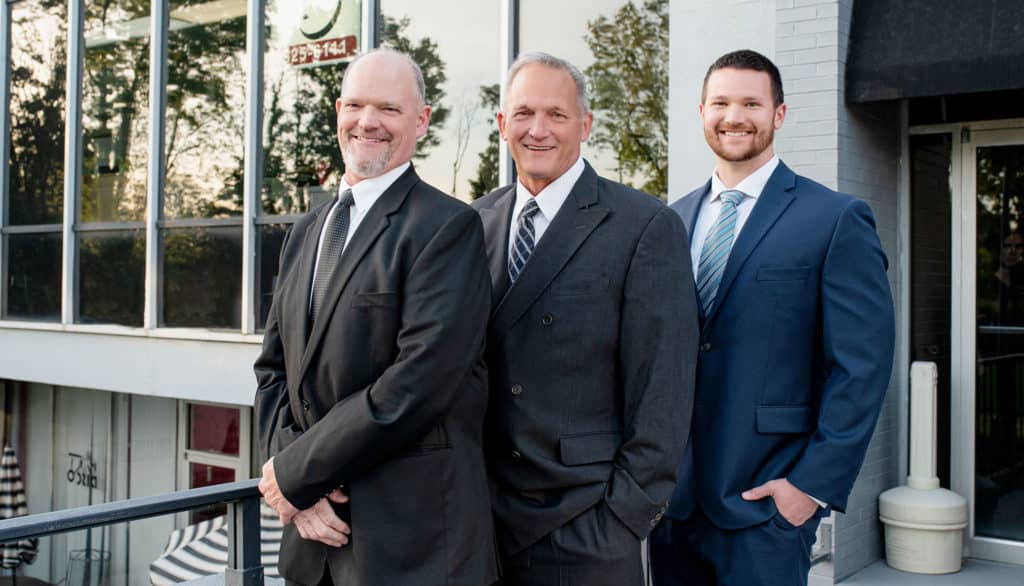 Three men in suits stand side by side, smiling, in front of a building with large glass windows. The man on the left has a beard and is wearing a black suit, the man in the middle is wearing a gray suit, and the man on the right is younger, wearing a navy blue suit with a light blue tie.
