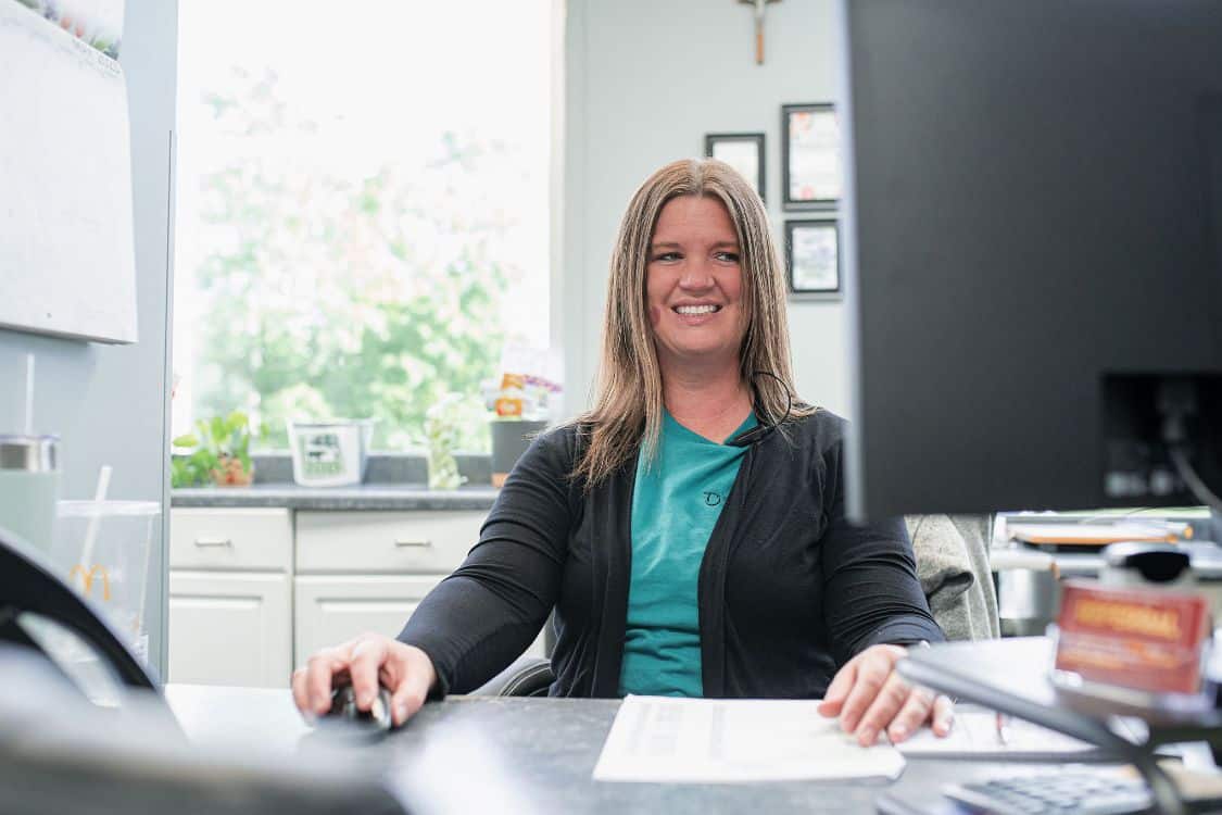 A dental office receptionist is smiling while working at her desk. She is using a computer mouse and looking at the screen, which is partially visible. The desk is organized with papers and a few personal items. The background includes a window with a view of greenery and framed certificates on the wall.