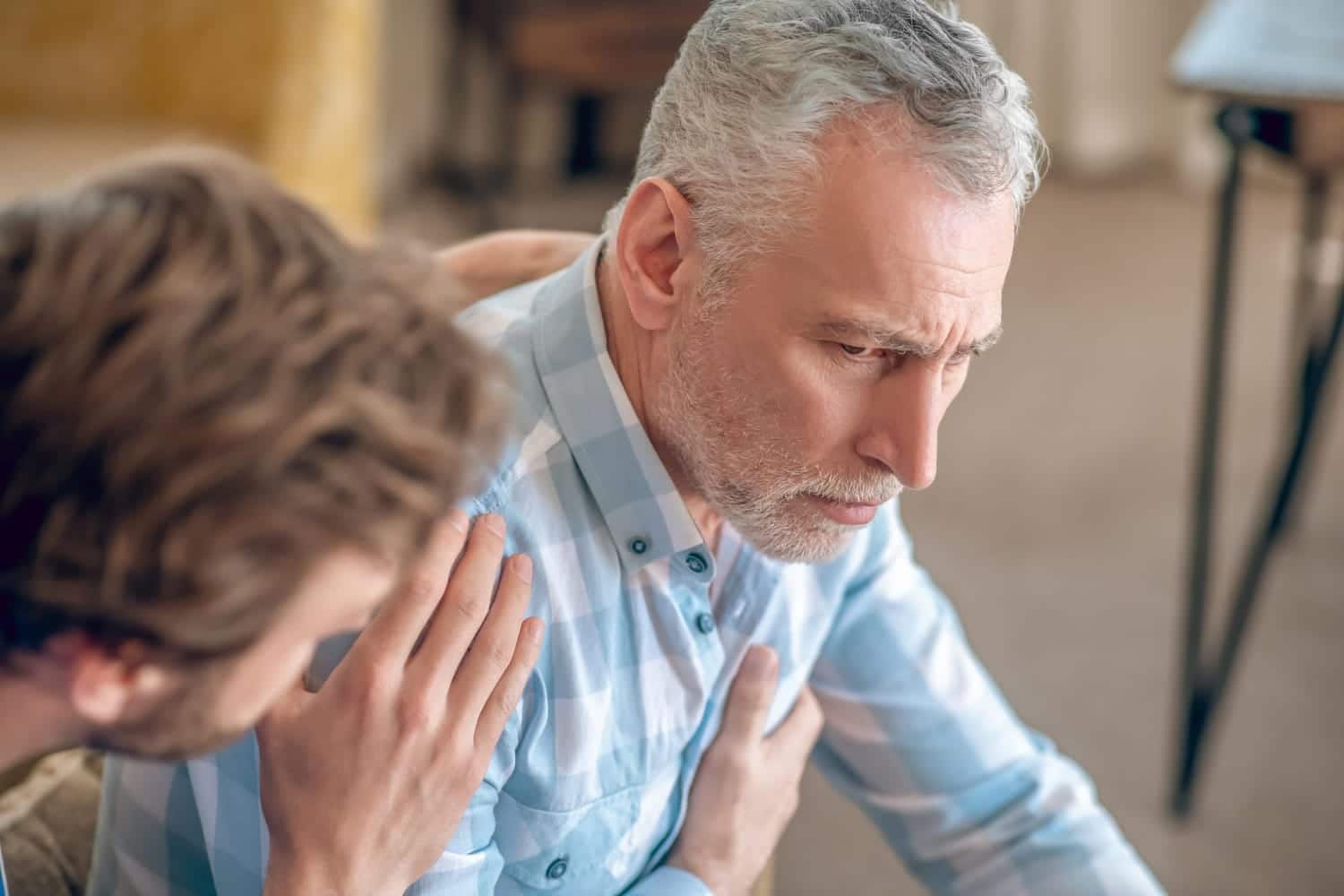 man with grey hair and beard holding his chest looks to be in pain